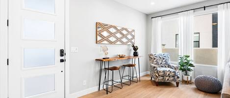 A modern living room with a wooden floor, a white door, a dining area with stools, a patterned armchair, and a large window with curtains.