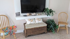 A living room with a flat-screen TV mounted on a white shelf, wicker furniture, and decorative items including seashells and a welcome sign. - Dream Vacation Pool and Waterfront Location (Saint James City)