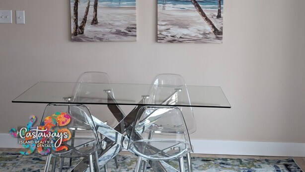 A dining area with a glass table and four chairs, two paintings of a beach scene on the wall, and a logo for Castaways Island Realty & Rentals. - Dream Vacation Pool and Waterfront Location (Saint James City)