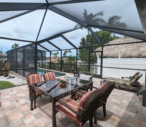 Une terrasse extérieure couverte, dotée d’une table en verre et de chaises en osier, qui offre une vue sur une piscine et une structure à toit de chaume.