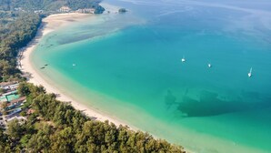 An aerial view of a coastal area with a sandy beach, clear turquoise waters, and several boats. There are buildings along the coastline, and the surrounding area is densely forested. - Dewa Phuket - Pool Villa (Sa Khu)
