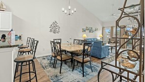 A dining area with a wooden table and chairs, a chandelier, and a view of the city through a large window. - Ocean Keyes 2242- 2BR/2BA-Close to Main St (North Myrtle Beach)