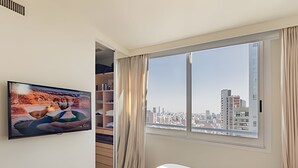 A hotel room with a bed, a TV mounted on the wall, a bookshelf, and a window with a city view. - Luxury and Spacious City Views Pool, Gym & Sauna (Buenos Aires)