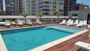 A rooftop pool with wooden decking, white lounge chairs, and a cityscape in the background. - Luxury and Spacious City Views Pool, Gym & Sauna (Buenos Aires)
