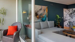 Un salon moderne avec un canapé d’angle gris, un fauteuil en osier avec un coussin rouge et une cloison en verre. Les murs sont ornés de tableaux abstraits et une plante se trouve sur la droite.