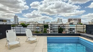 Une piscine sur le toit, agrémentée de deux chaises longues blanches, avec une vue sur les toits de la ville en arrière-plan et un ciel dégagé.