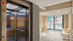 Un ascenseur doté d’une porte en verre, un espace d’attente avec des chaises et une vue sur la rue grâce à de grandes fenêtres.