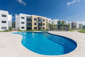 A swimming pool with a curved edge, surrounded by modern buildings with balconies and a clear sky. - Pool View and 7 Minutes from the Beach (Punta Cana)
