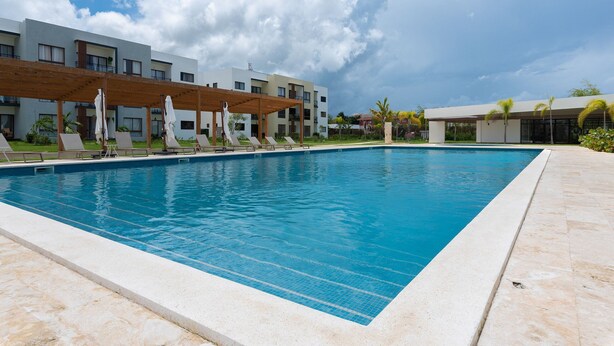 A rectangular swimming pool with a tiled edge, surrounded by lounge chairs and umbrellas under a wooden pergola.