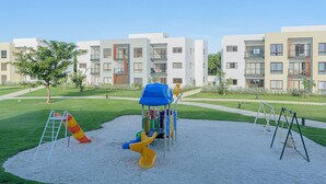 A playground with slides and swings, surrounded by a well-maintained lawn and modern apartment buildings in the background.
