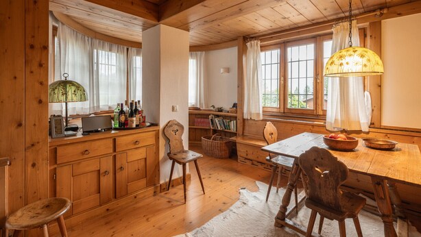 Un interior rústico de madera con una mesa de comedor de madera, sillas, una lámpara y una ventana con cortinas.