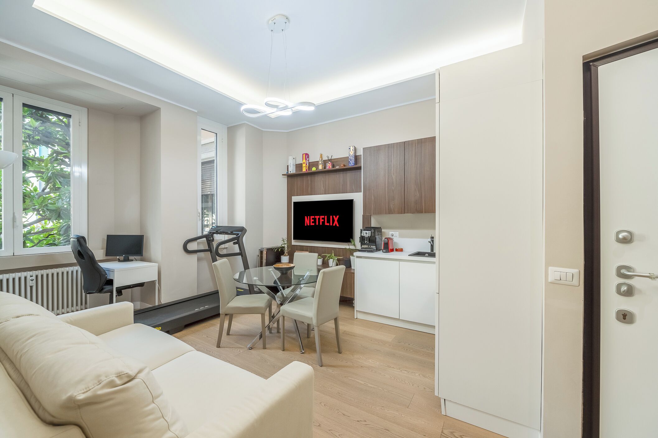 Een moderne woonkamer met een bank, een eethoek met een glazen tafel, een loopband en een tv waarop 'NETFLIX' wordt weergegeven.