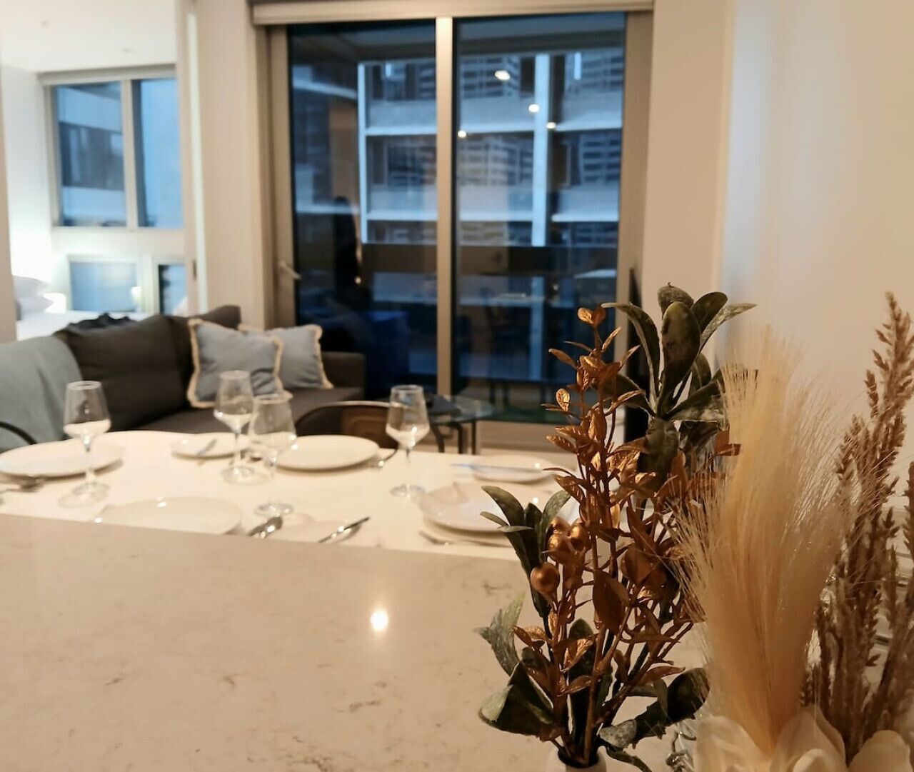 A dining area with a table set for four, a sofa in the background, and a view of a building through large windows.