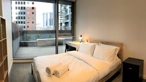 Ein Hotelzimmer mit großem Fenster, einem Bett mit weißen Laken, einem Nachttisch mit Lampe und einem kleinen schwarzen Schrank.