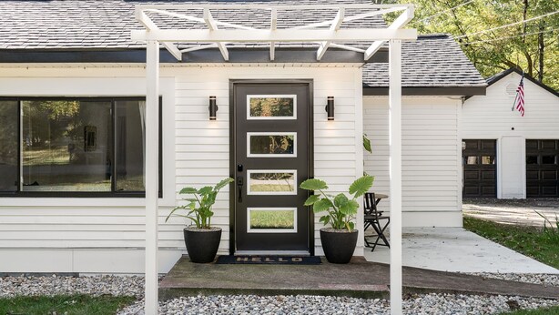 Una casa moderna con fachada blanca, puerta principal oscura con dos ventanas y una pérgola sobre la entrada. Hay dos macetas con plantas a cada lado de la puerta.
