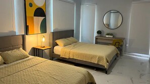 A bedroom with two beds, a bedside table with a lamp, a desk with a chair, a round mirror, and abstract wall art.