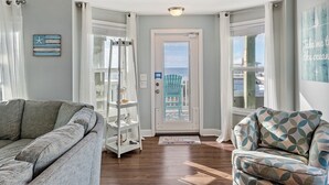 A living room with a grey sofa, a patterned armchair, a white shelving unit, and a door with a view of the city. - At Last - Oceanfront - 5 Bed, 4 Bath -  Pool, Hot Tub, Elevator & Backyard (North Topsail Beach)