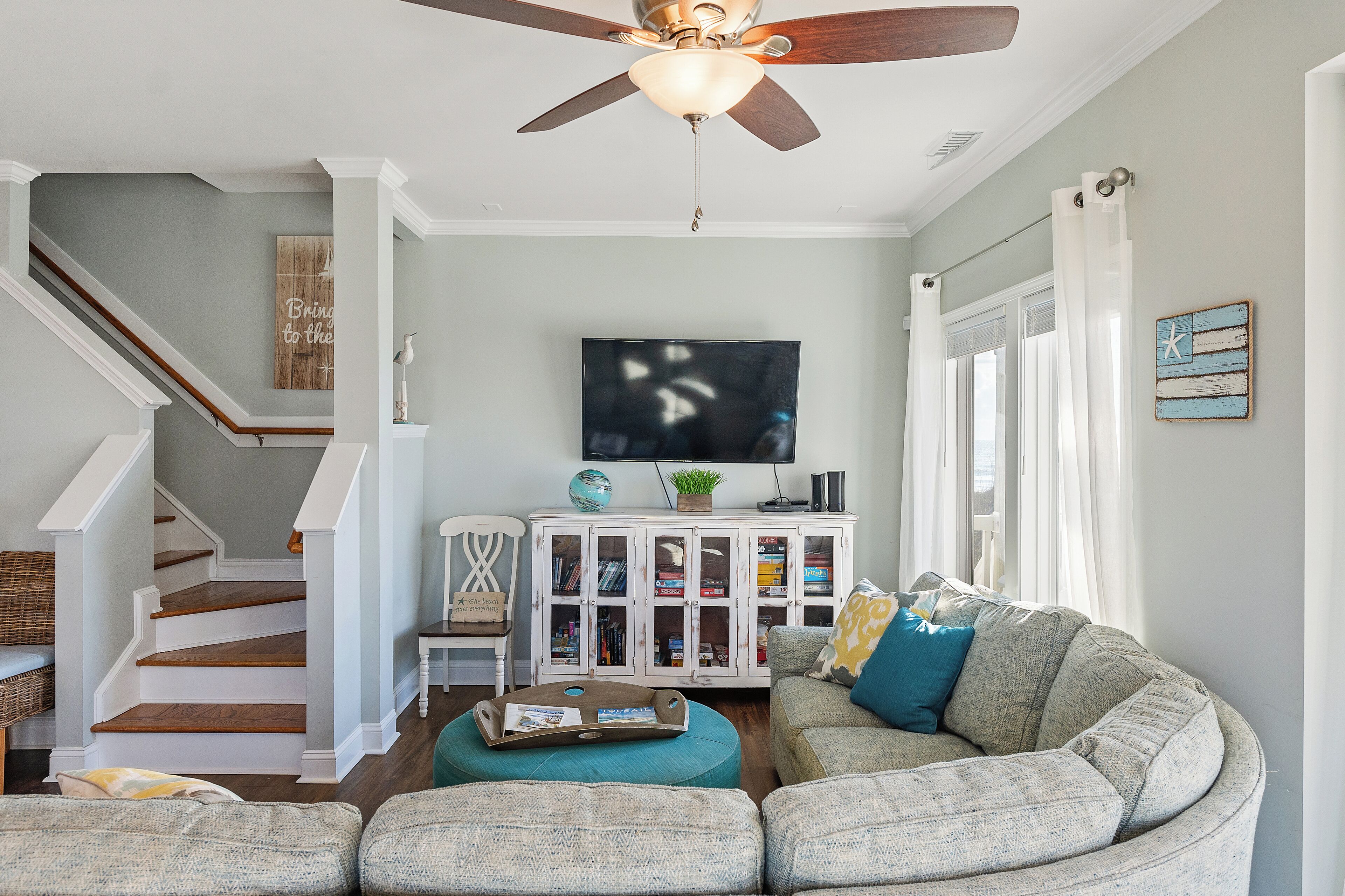 Una sala de estar con escalera, ventilador de techo, televisor y un aparador con libros y objetos decorativos.