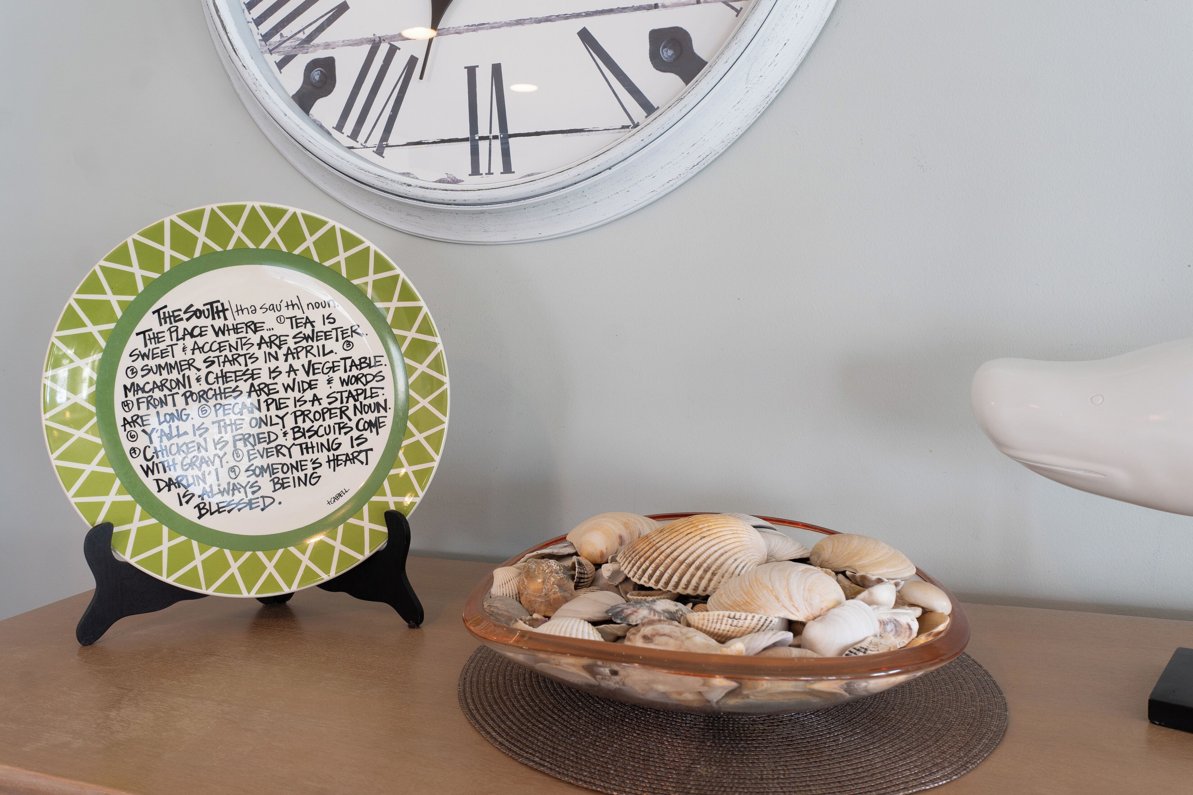 A clock with Roman numerals, a decorative plate with text, and a bowl of seashells on a table.