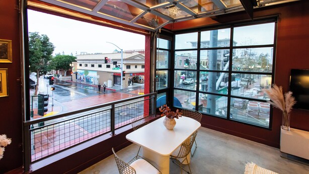 A modern interior with a large window overlooking a busy street, a white table with chairs, and a vase with dried plants. - Studio loft with garage door with AC located in the heart of DTSA (Santa Ana)