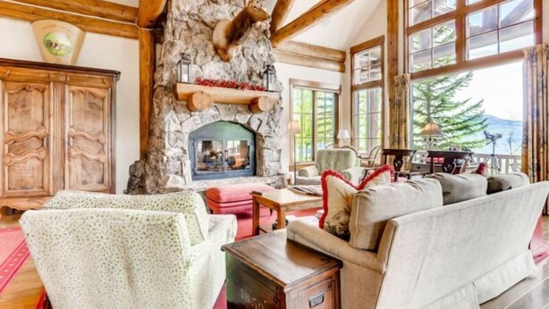 A cozy living room with a stone fireplace, wooden beams, and large windows offering a view of the outdoors.