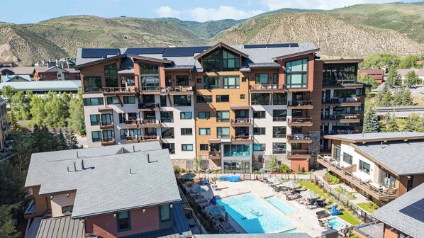 A multi-story hotel with balconies, a swimming pool, and a mountainous backdrop.