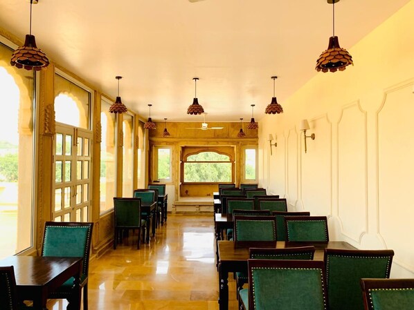 A dining hall with rows of tables and chairs, large windows, and hanging pendant lights. - Hotel Dev Garh (Jaisalmer)