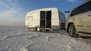Valkoinen matkailuauto on pysäköitynä beigenvärisen maasturin viereen lumiselle alustalle.
