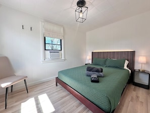 A bedroom with a bed, bedside tables, a chair, a window with blinds, and a ceiling light fixture. - Peaceful Escape • Hot Tub • Fire Pit • Views (Swan Lake)