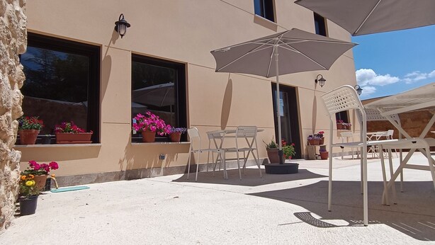 A building with a beige exterior, large windows, outdoor seating area with white chairs and tables, and potted plants.