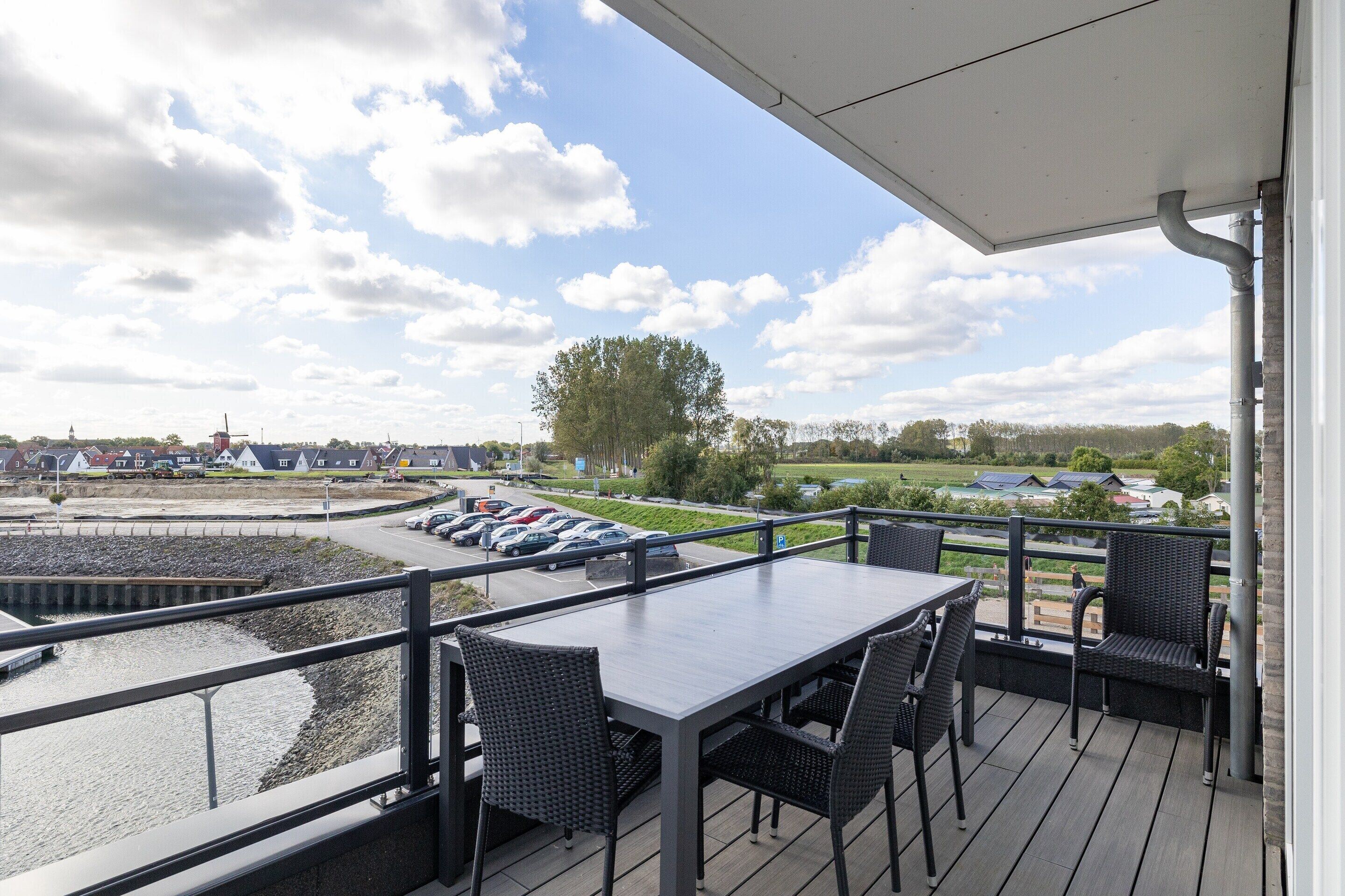 A balcony with a table and chairs overlooking a parking area and a body of water.