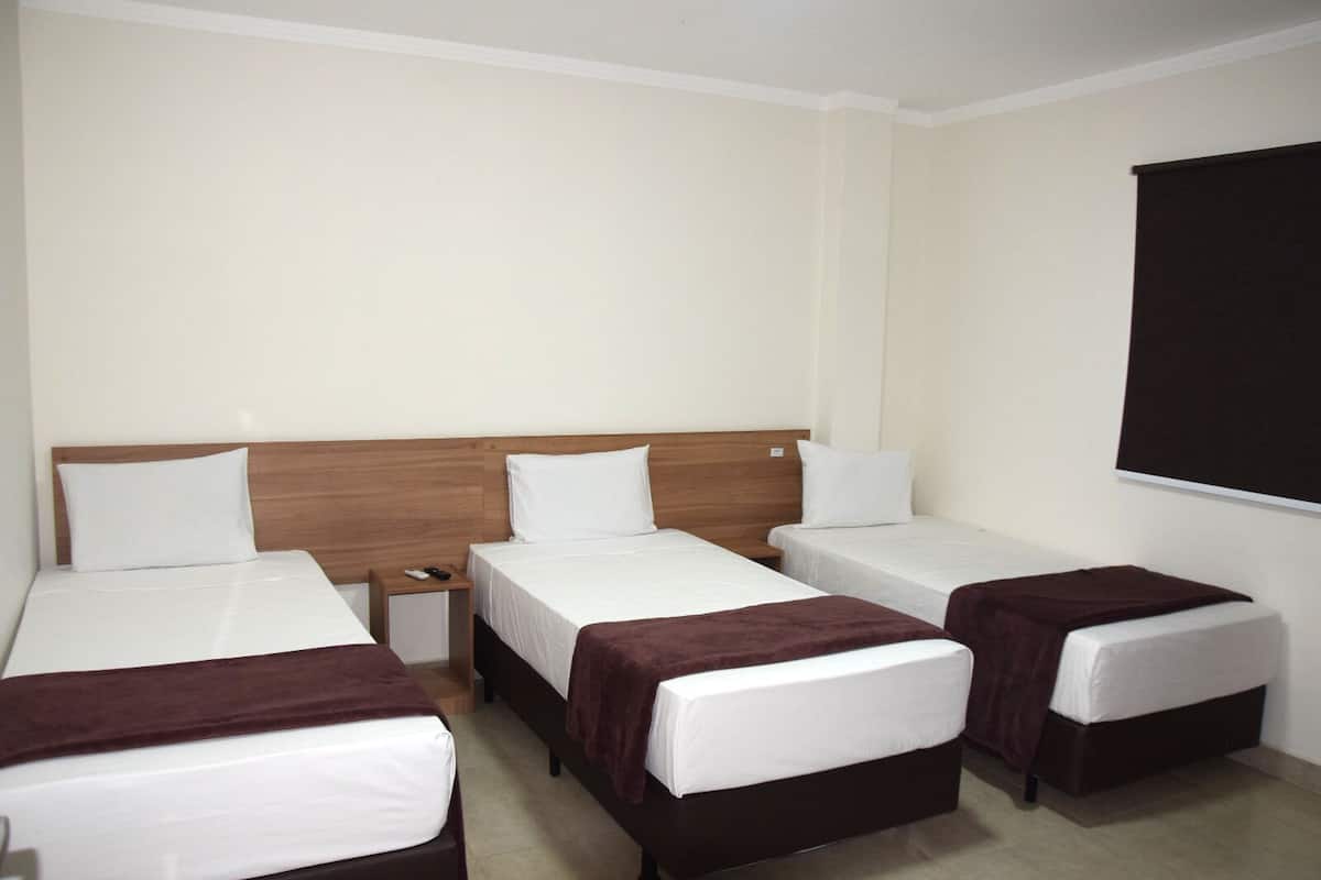 A room with three single beds, each with a wooden headboard and white bedding. There is a small wooden nightstand with a remote control on it.