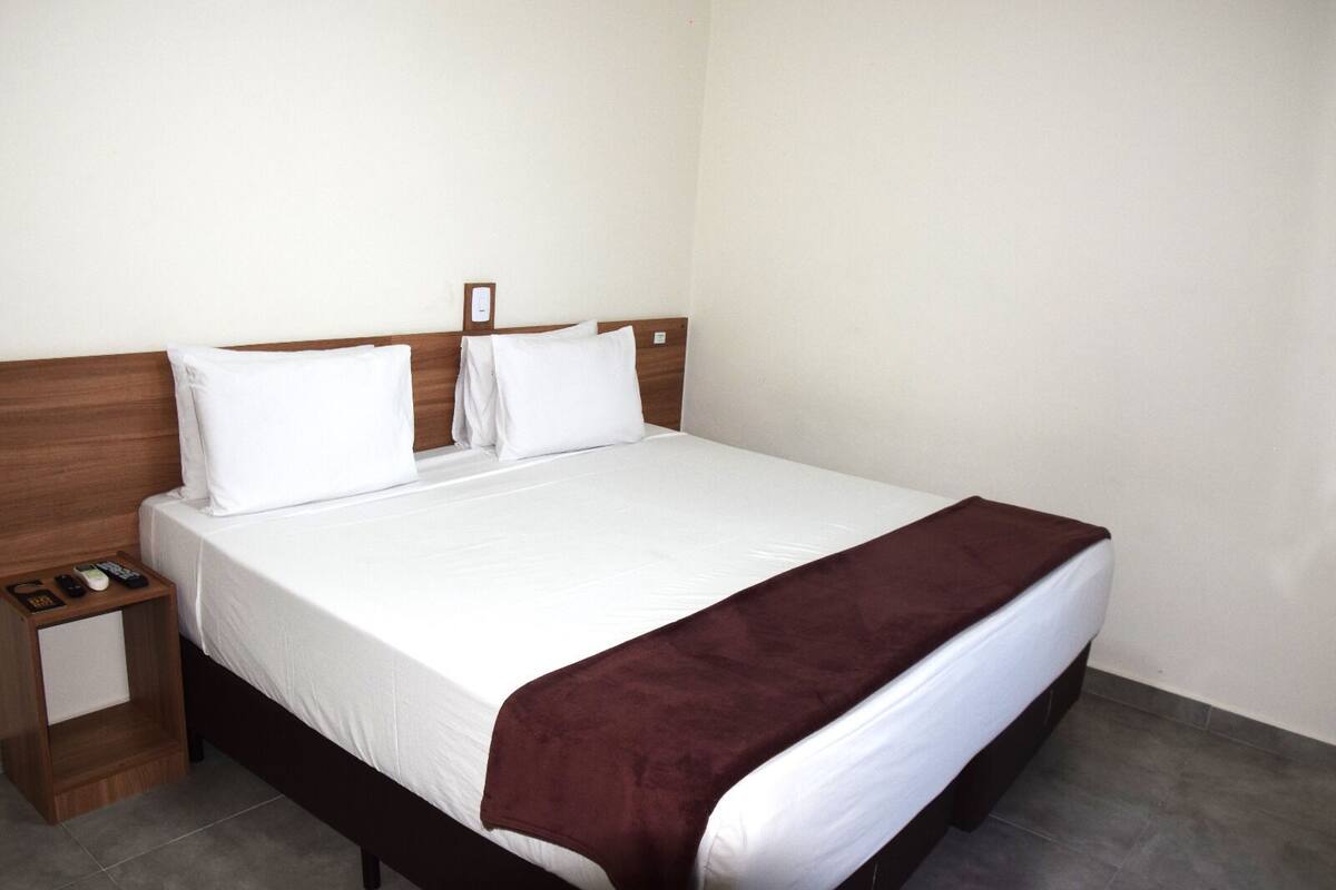A neatly made bed with white linens and a brown throw blanket, a wooden headboard, and a small bedside table with a remote control and a book.