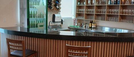 A bar area with wooden bar stools, a refrigerator stocked with bottles, and shelves with glasses and liquor bottles.