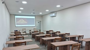 A classroom with wooden desks and chairs, a projector screen, and a ceiling-mounted projector. - HOTEL INTERNACIONAL GUARULHOS (Guarulhos)