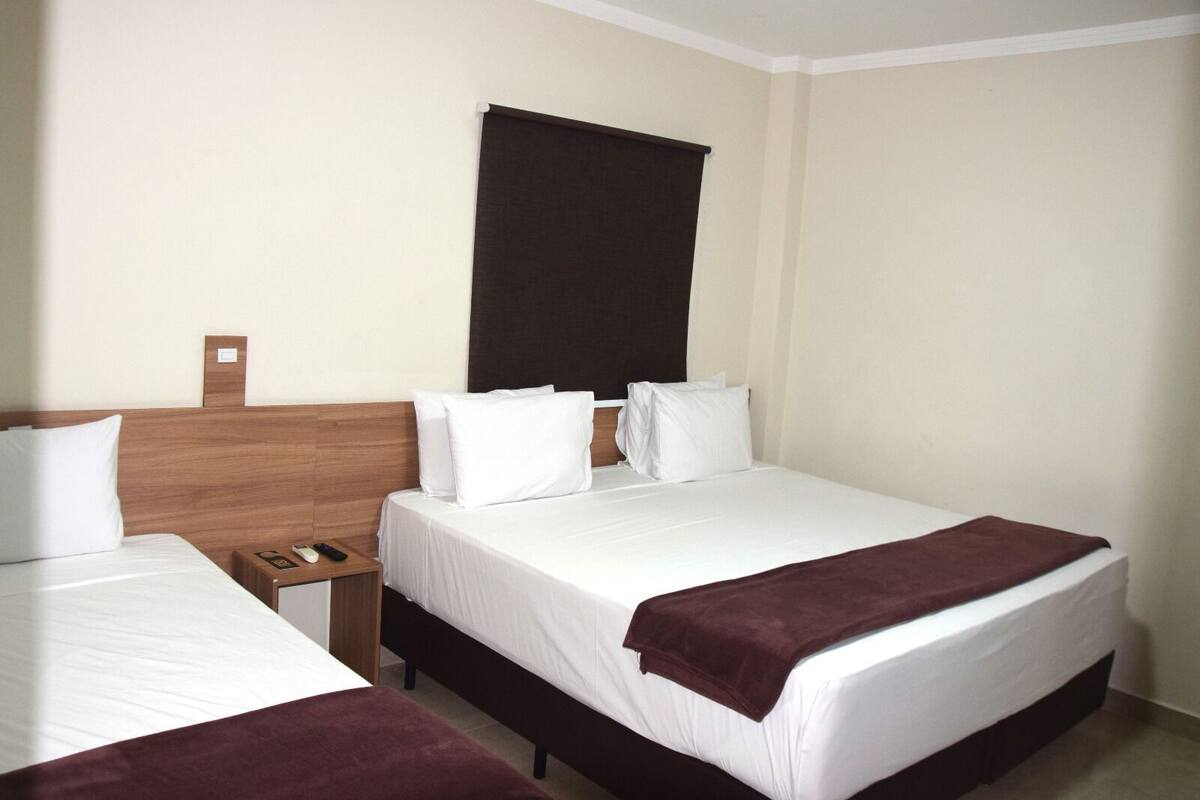 A hotel room with two beds, a wooden headboard, a dark-colored window blind, and a small bedside table with a remote control.