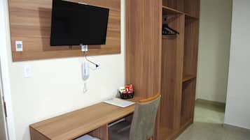 Una stanza con una scrivania in legno, una sedia, una televisione a muro, un armadio e un tavolino con una tazza e una confezione.