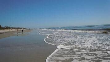 A beach with waves gently lapping onto the shore, a clear sky, and a few people walking along the beach.