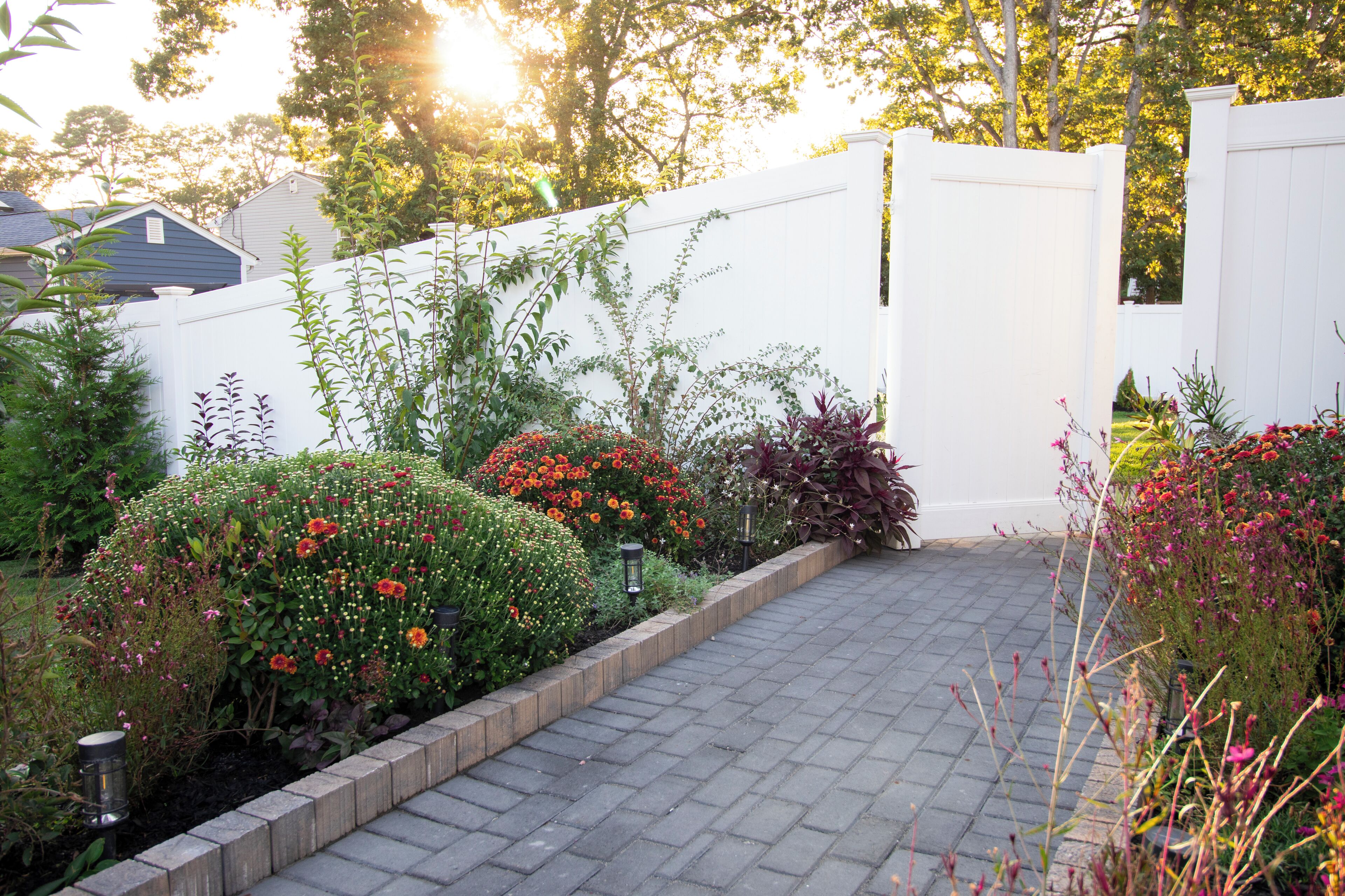 A paved pathway leading to a white fence, surrounded by various flowering plants and shrubs.