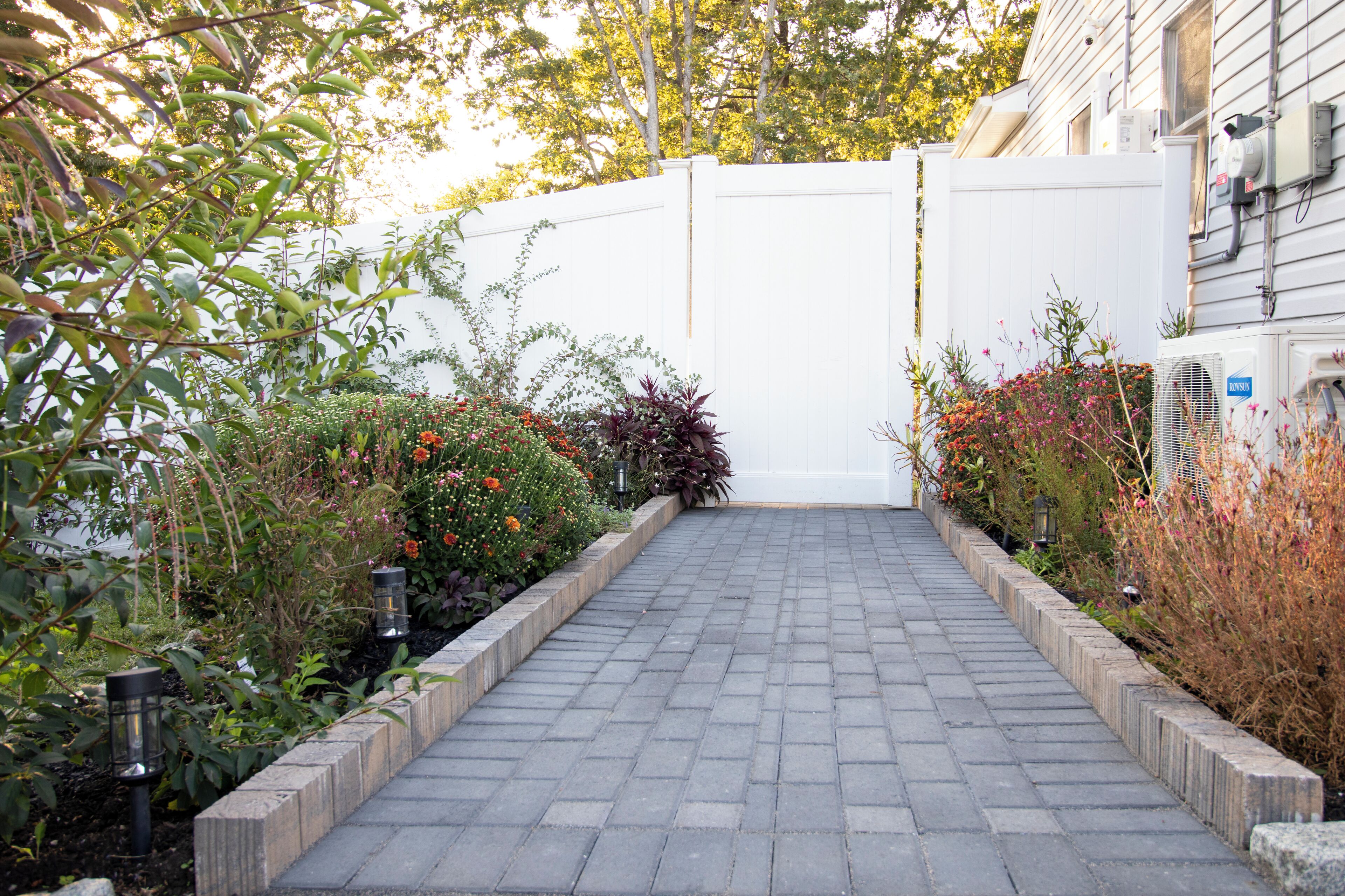 Un chemin pavé mène à un portail blanc, qui est entouré de différentes plantes et fleurs.