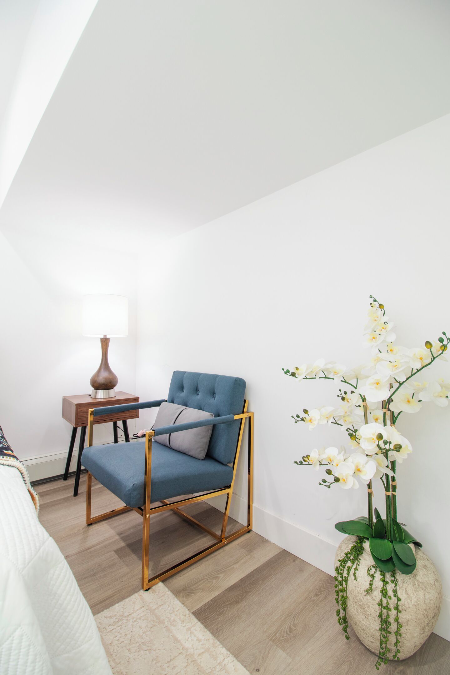 Quarto com uma poltrona azul, uma mesa de cabeceira de madeira com uma luminária, um vaso com orquídeas brancas e uma cama com cobre branca.