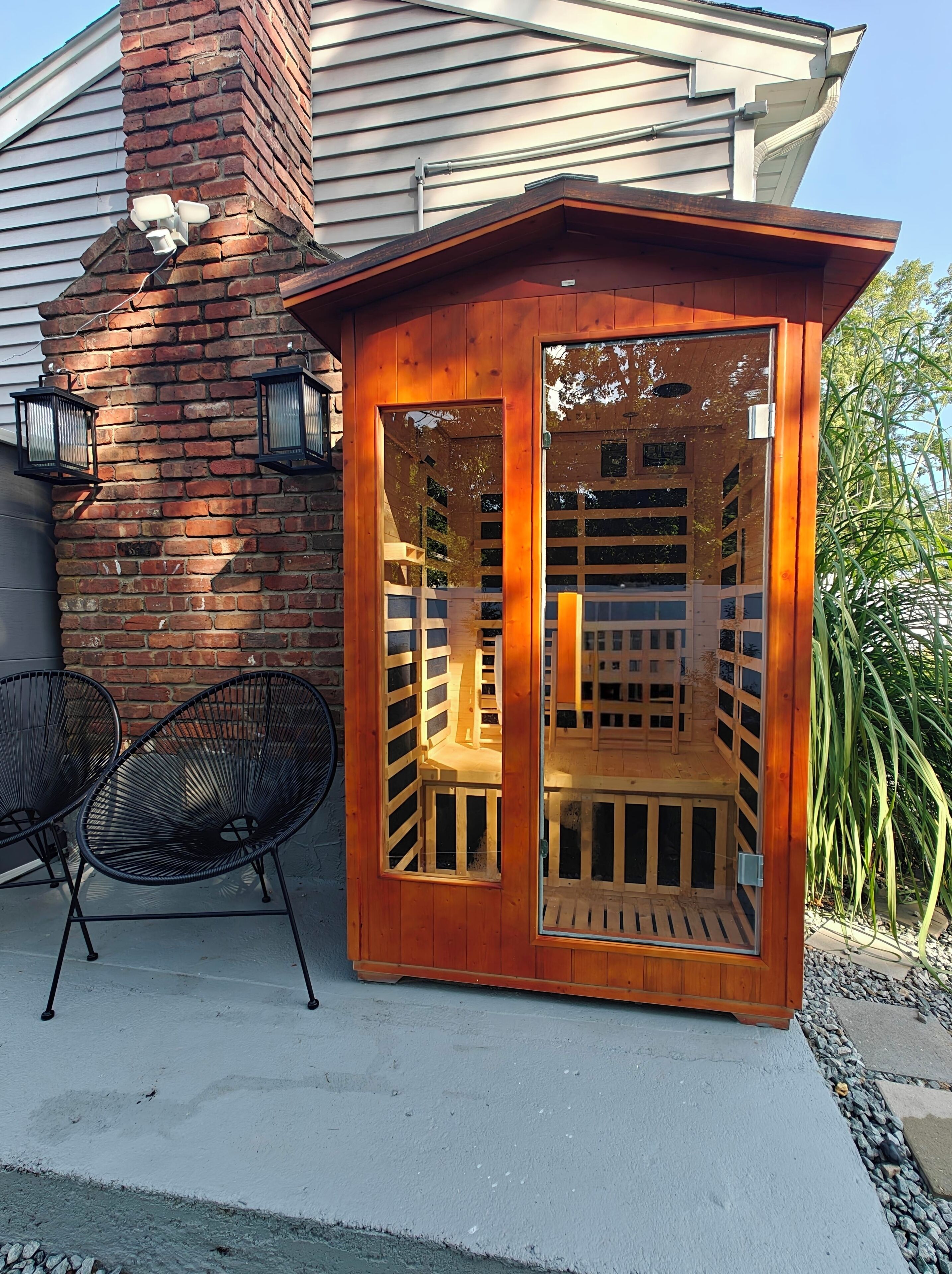 A wooden outdoor seating area with a bench and two chairs, a brick wall, and a window.