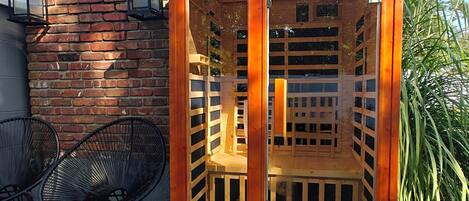 A wooden outdoor seating area with a bench and two chairs, a brick wall, and a window.
