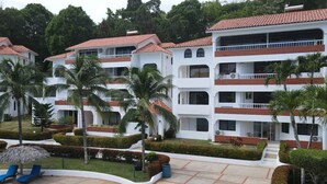 A multi-story hotel with white exterior walls and red-tiled roofs, featuring balconies, a swimming pool, and well-maintained gardens.