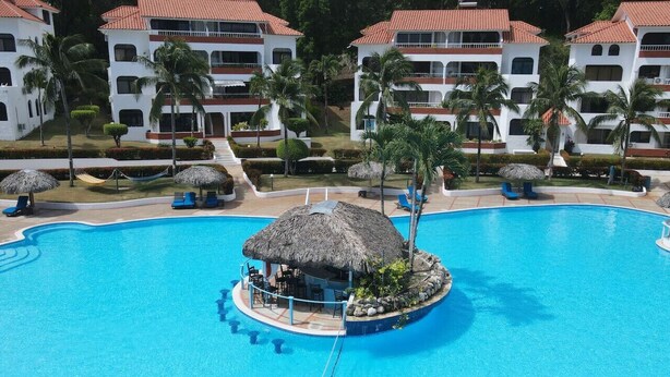 A large swimming pool with a thatched-roof structure in the center, surrounded by palm trees and lounge chairs.