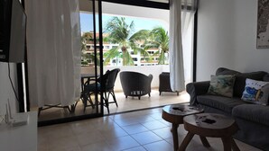A living room with a sofa, two wooden side tables, and a dining area with chairs and a table. A large window offers a view of palm trees and buildings.