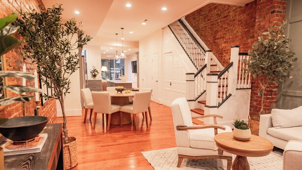 A modern living room with a wooden floor, a dining area with a table and chairs, a staircase, and a brick wall.