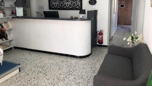 Zona de recepción con un escritorio blanco, estanterías con libros y objetos decorativos, una obra de arte blanco y negro, y un extintor.