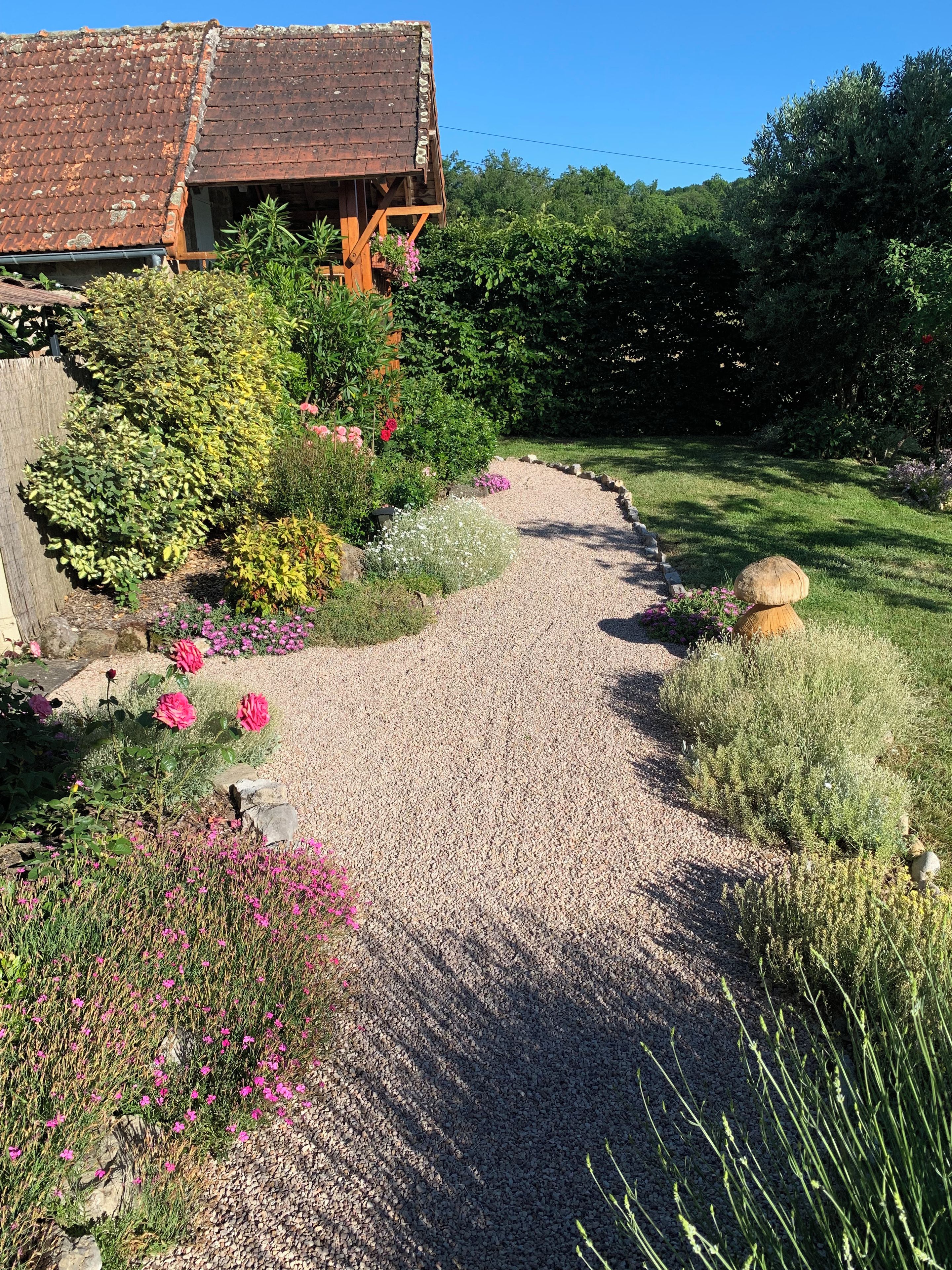 A gravel pathway leading to a rustic wooden structure with a tiled roof, surrounded by lush greenery and flowering plants.