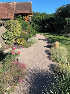 A gravel pathway leading to a rustic wooden structure with a tiled roof, surrounded by lush greenery and flowering plants.
